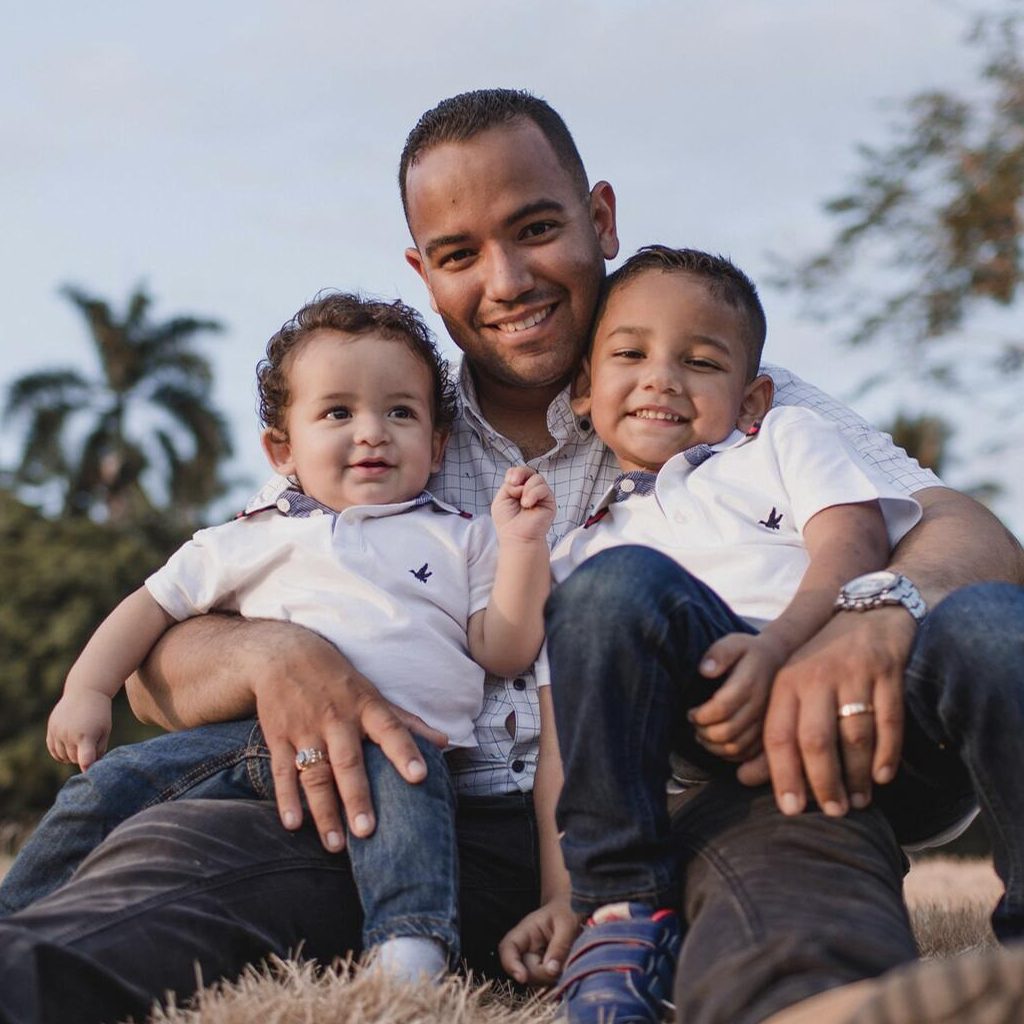 smiling man sitting on the ground with his two young children sitting on his lap