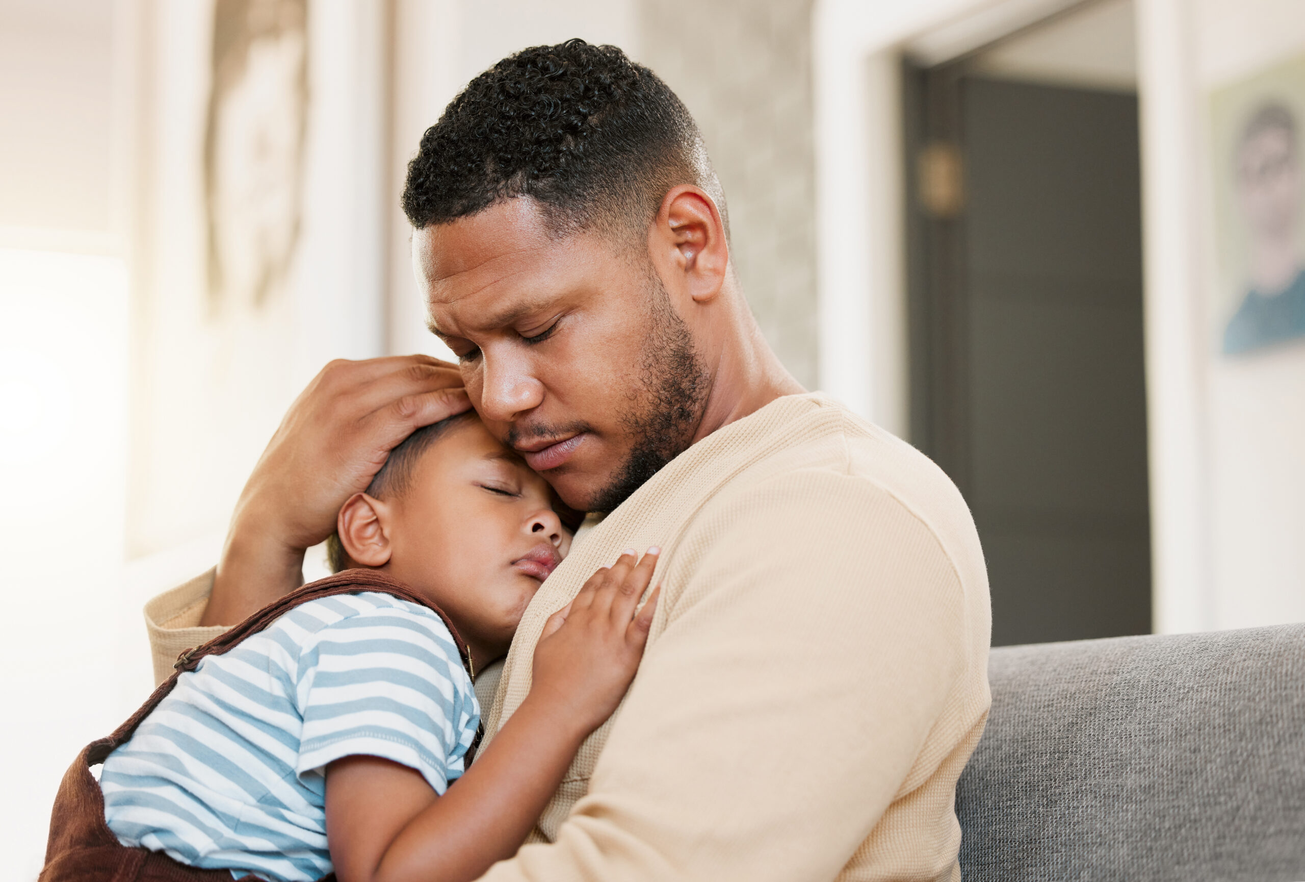 Sleeping and tired child with caring father holding his sleepy son on a home lounge sofa. Worried dad hugging his young boy resting on a living room family couch indoors feeling stress and worry